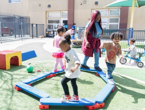 A Head Start educator walks with young children on beams set in a circle on grass outside while other children play in the background