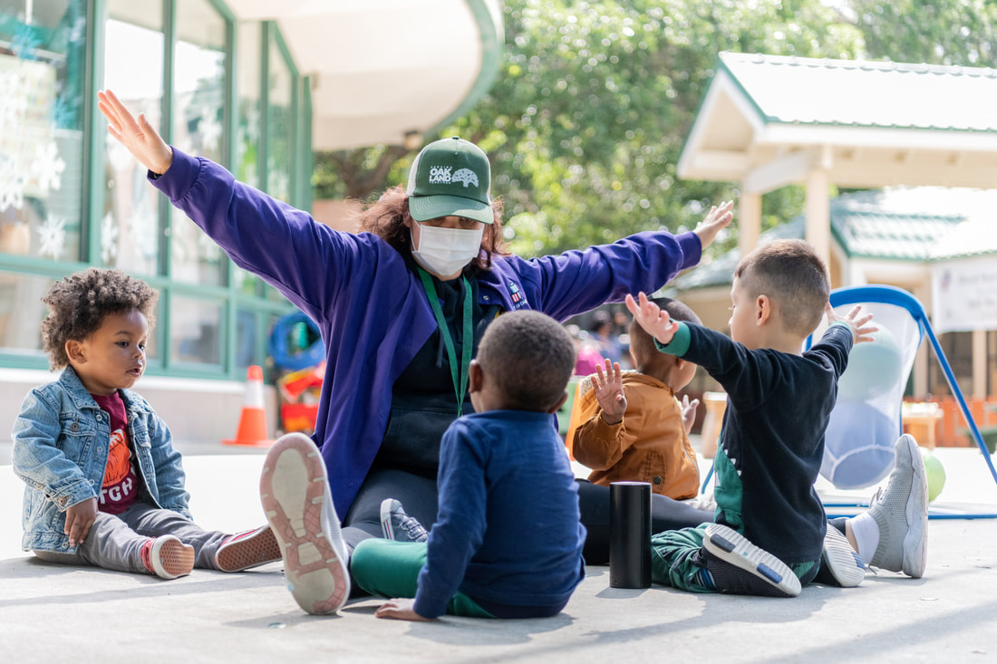 An educator practices yoga outside with several young children, stretching her arms wide while seated
