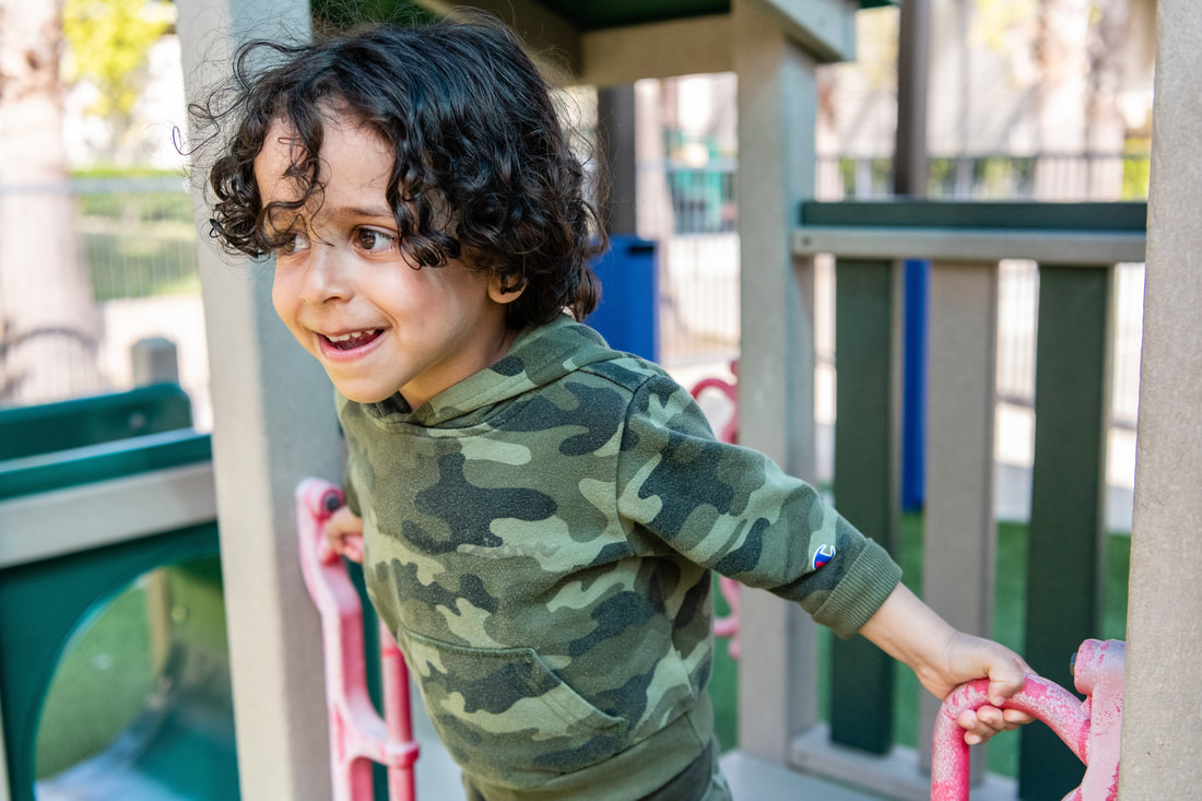 A young child with curly hair smiles while leaning forward in a play structure