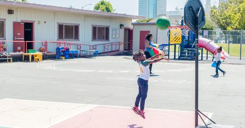 Young girl with braids plays outside in a schoolyard, jumping up to toss a ball through a basketball hoop 