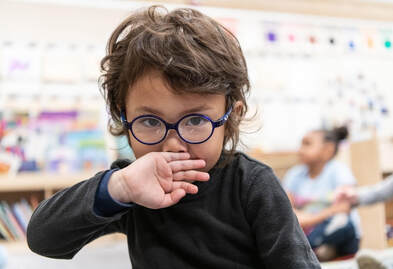A young child with brown hair wears round, black glasses and holds their hand, palm out, in front of their mouth