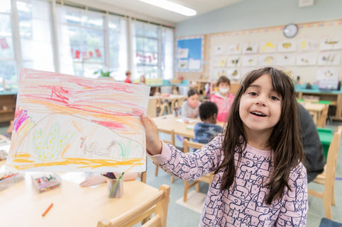A young child in a pink shirt smiles while showing a colorful drawing