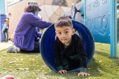 A young child crawls out of a blue fabric tunnel onto the grass