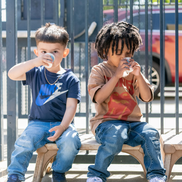Two young children drink from paper cups while sitting on a bench outside